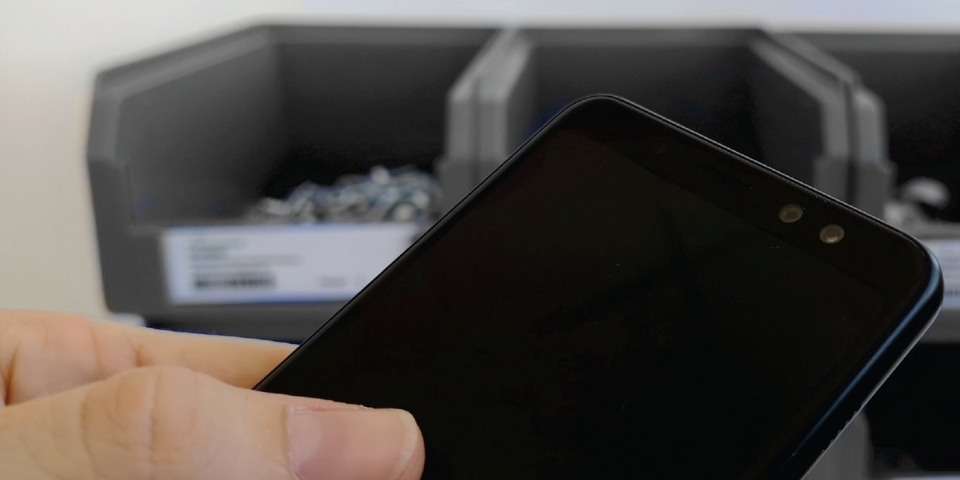 Im Vordergrund eine Hand, die ein schwarzes Smartphone mit ausgeschaltetem Bildschirm hält. Im Hintergrund sind drei graue Aufbewahrungsbehälter mit kleinen Gegenständen darin auf einer weißen Oberfläche platziert.